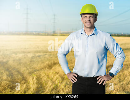 Giovani collare blu lavoratore o ingegnere indossa nel casco giallo si erge nel campo con un ampio sorriso. mezza lunghezza verticale. Foto Stock