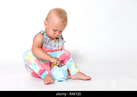 Raffreddare il toddler ragazza seduta sul pavimento è di gettare una moneta nell salvataggio salvadanaio. Il tutto è su sfondo bianco. Orizzontalmente. Foto Stock