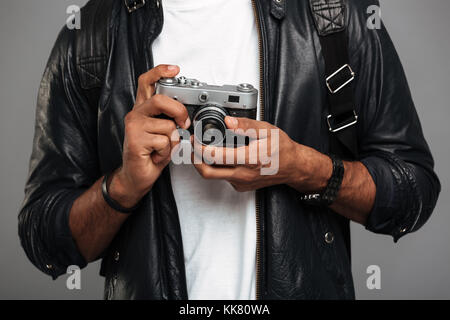 Close up di un giovane maschio africano fotografo in giacca di pelle tenendo la fotocamera retrò isolate su uno sfondo grigio Foto Stock