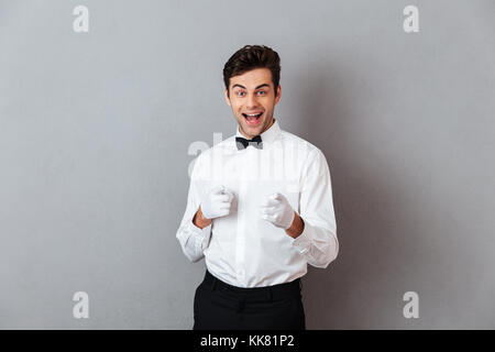 Ritratto di un allegro giovane maschio cameriere vestito in unifrom che puntano a due dita fino alla telecamera isolate su uno sfondo grigio Foto Stock