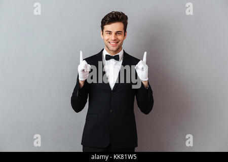 Ritratto di un sorriso amichevole uomo vestito in smoking e guanti che puntano a due dita verso l'alto isolato su sfondo grigio Foto Stock