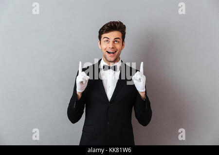 Ritratto di un allegro giovane uomo vestito in smoking e guanti che puntano a due dita verso l'alto isolato su sfondo grigio Foto Stock