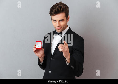 Ritratto di un bel affascinante uomo vestito in smoking tenendo aperta la casella con un anello di fidanzamento e puntare il dito in telecamera isolate su backgr grigio Foto Stock