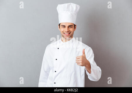 Immagine di felice giovane cuoco in uniforme isolato in piedi oltre il muro grigio Sfondo. Guardando la telecamera che mostra Thumbs up. Foto Stock