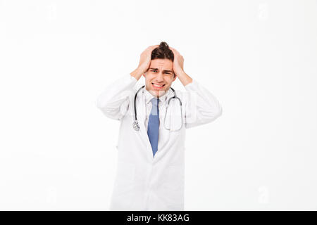 Ritratto di un frustrato giovane maschio medico con stetoscopio vestito in uniforme in piedi con le braccia alla sua testa e guardando la telecamera isolate su whit Foto Stock