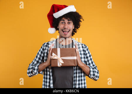 Ritratto di un felice allegro african uomo vestito di natale hat azienda confezione regalo e guardando la telecamera isolate su sfondo arancione Foto Stock