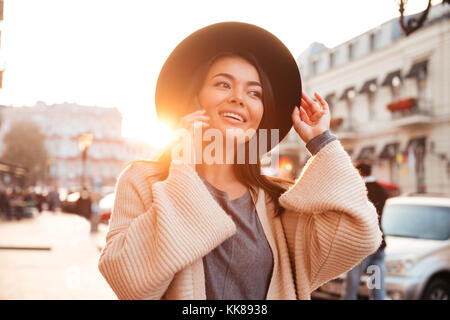 Incantevole donna asiatica regolando il suo cappello nero mentre parlano al telefono mobile sulla strada di città Foto Stock