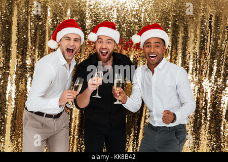 Gruppo di felice allegro multirazziale amici in cappelli di Natale celebrare durante il riposo e bere champagne isolate su golden sfondo lucido Foto Stock