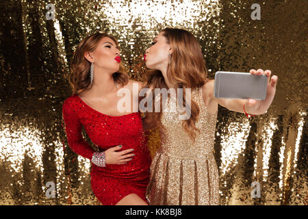 Ritratto di due attraenti le giovani donne in abiti scintillanti prendendo un selfie mentre in piedi e in posa isolate su golden sfondo lucido Foto Stock