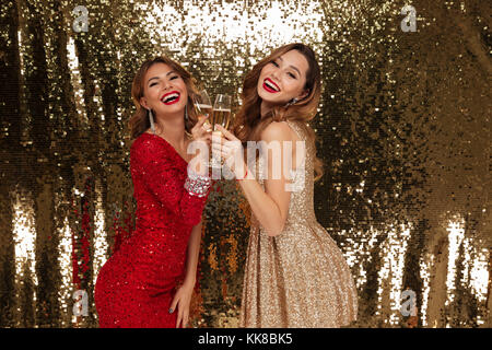 Ritratto di due belle ragazze sorridenti in abiti scintillanti holding bicchieri di champagne mentre in piedi e celebrando isolate su golden backgro lucido Foto Stock