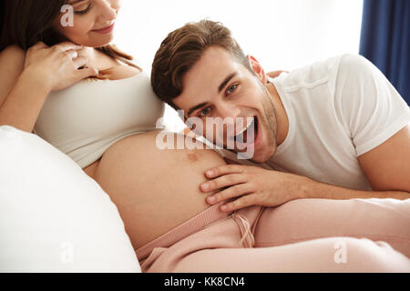 Felice giovane uomo che ascolta sua moglie incinta la pancia mentre giaceva insieme sul letto in ambienti interni Foto Stock