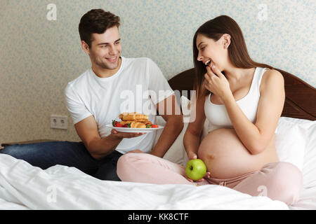 Ritratto di un giovane eccitato incinta giovane avente una sana colazione insieme seduti a letto Foto Stock