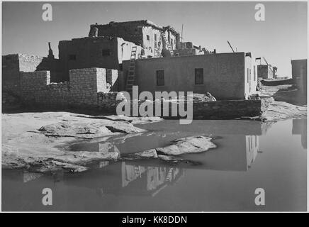 Fotografia in bianco e nero, piena vista laterale di adobe house con acqua in primo piano, con la dizione "Acoma Pueblo Pietra Miliare Storica Nazionale, Nuovo Messico', di Ansel Adams, da fotografie di parchi e monumenti nazionali, Nuovo Messico, Stati Uniti, 1941. Foto Stock