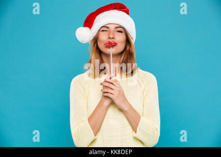 Close-up di giovane donna attraente in Santa's hat tenendo le labbra di carta su un bastone mentre lampeggia un occhio, isolato su sfondo blu Foto Stock