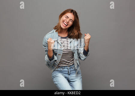 Ritratto di un gioioso felice ragazza adolescente vestito in camicia denim celebra il successo mentre balli isolate su uno sfondo grigio Foto Stock