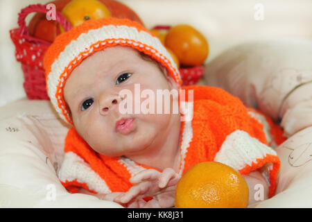 Baby di inviare aria kiss. Ritratto di neonato in arancione costume di maglia su coltre bianca e il cestello con le arance e zucca dietro di lui di inviare un Foto Stock