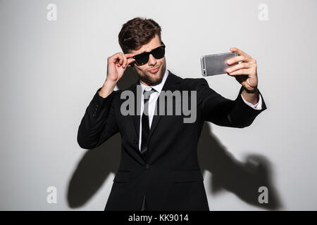 Giocoso macho in black classic tuta e occhiali da sole prendendo selfie sullo smartphone, isolato su sfondo bianco Foto Stock