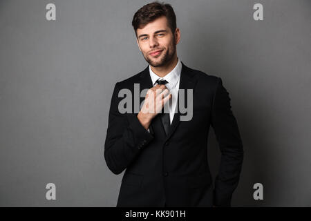 Giovane uomo attraente in abito classico si raddrizza la cravatta, guardando la telecamera, isolata su uno sfondo grigio Foto Stock