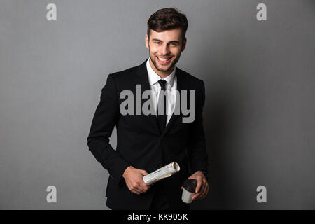 Giovani sorridente uomo barbuto in convenzionale tenendo il giornale e tazza di caffè, guardando la telecamera, isolata su uno sfondo grigio Foto Stock