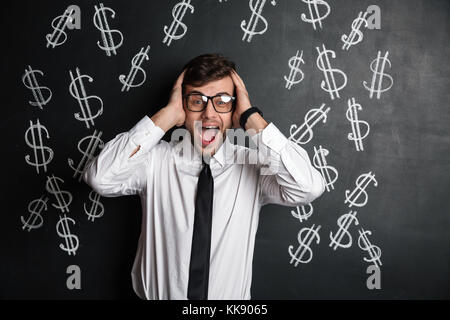 Overjoyed giovane uomo barbuto in bicchieri tenendo la sua testa, guardando la telecamera, isolate su lavagna con simbolo di dollari Foto Stock