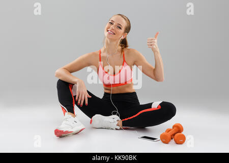 Ritratto di un felice piuttosto sportsgirl ascoltare musica con auricolari durante la seduta sul pavimento e mostra Thumbs up gesto isolato grigio su ba Foto Stock