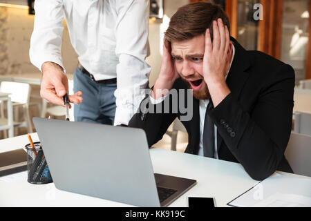 Giovani scioccato barbuto imprenditore nel convenzionale con la bocca aperta tenendo la sua testa, mentre il suo partner puntando con penna su uno schermo del notebook in ufficio Foto Stock