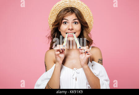 Ritratto di un bel colore marrone ragazza dai capelli in estate hat mangiando caramelle di cotone e guardando la telecamera isolate su sfondo rosa Foto Stock