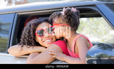 Divertimento estivo! Le fanciulle felici o sorelle con a forma di cuore ad occhiali da sole nella finestra auto. Giovane ragazza baci ragazza più vecchi sulla guancia in celebrazione dell amore, estate Foto Stock