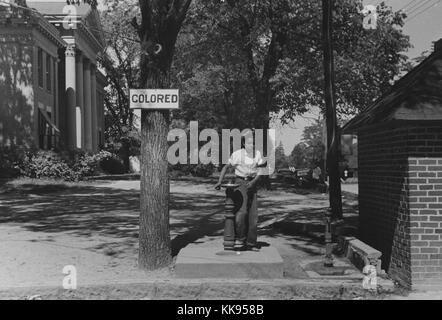 Una fotografia di un Africano bambino americano con una fontana di acqua, l'albero accanto alla fontana ha un segno attaccato che recita "colorato" al fine di imporre Jim Crow leggi, fontana di acqua è parte del County Courthouse proprietà, Halifax, North Carolina, 1938. Dalla Biblioteca Pubblica di New York. Foto Stock