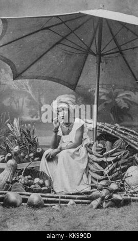 Un ritratto fotografico di una donna seduta su una sedia tra vari frutti tropicali e verdure, la donna indossa una lunghezza pianale abito bianco e una luce colorata testa wrap, lei sta fumando un sigaro e posizionato sotto un grande ombrellone, platani, noci di cocco, ananas e zucche di grandi dimensioni può essere visto, la fotografia è stata allestita in un studio, un fondale dipinto raffigurante una scena tropicale è visibile dietro di lei, Brasile, 1874. Dalla Biblioteca Pubblica di New York. Foto Stock