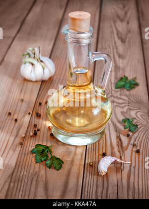 Un decanter di vetro con olio vegetale su un sfondo di legno circondato dal verde, aglio e pepe è chiusa con un tappo di sughero Foto Stock