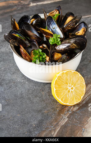 Cozze con erbe aromatiche in una ciotola bianca con limone. Il pesce e il cibo presso la riva del mare francese. sfondo scuro Foto Stock