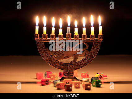 Hanukkah menorah con candele accese si siede sul tavolo; le luci si riflettono nella finestra scuro dietro di loro; dreidels sparsi sul tavolo di fronte o Foto Stock