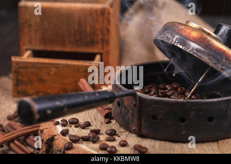 Il dispositivo per la tostatura i chicchi di caffè, un vecchio macinino a mano. fumoso caffè arrosto su un supporto di legno. studio di illuminazione. Foto Stock