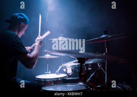 Il batterista in un cappuccio e cuffie suona la batteria in un concerto sotto la luce blu in un fumo Foto Stock