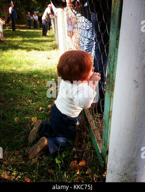 Carino piccolo bambino con zenzero capelli guardando attraverso la recinzione in piedi sulle ginocchia il solo tra persone adulte intorno a. Foto Stock