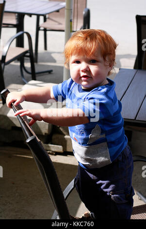 Fashioned ginger toddler ragazzo con gli occhi blu di stare sulla sedia di vimini in aperto estate cafe' all'aperto. Foto Stock