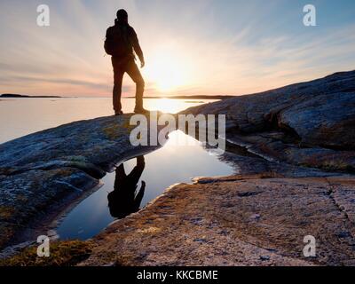 Tall backpacker guarda chiara primavera soleggiata alba sopra il livello del mare. Escursionista con zaino stare sulla spiaggia rocciosa e la sua figura è specchiata in acqua piscina. Escursionista Foto Stock