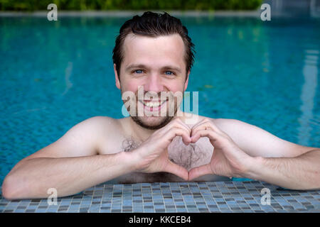 Bello sorridente giovane uomo caucasico rende il cuore usando le dita, le mani durante il nuoto in piscina. Foto Stock