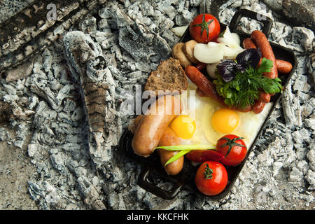 Salsicce affumicate con pomodori e uova giacciono su carbone. il piatto è cotto e affumicato su carbone di legna Foto Stock