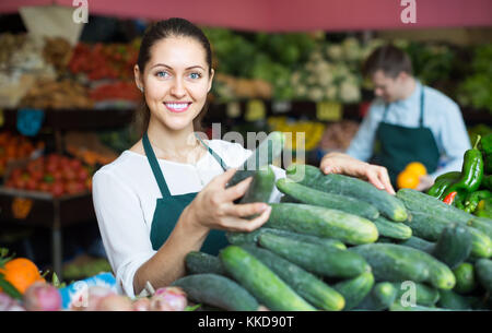 Roba sorridente nel grembiule verde vendita cetriolo al marketplace Foto Stock