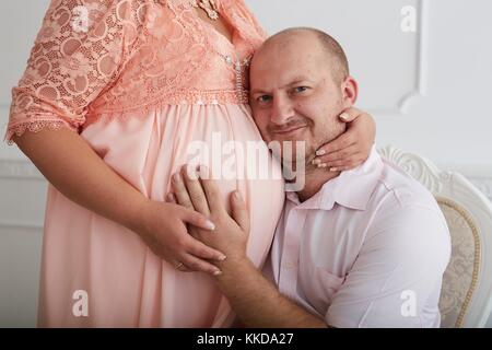 L'uomo abbracciando ventre della donna incinta in abito di corallo sul lusso decorate muro bianco sullo sfondo. Foto Stock