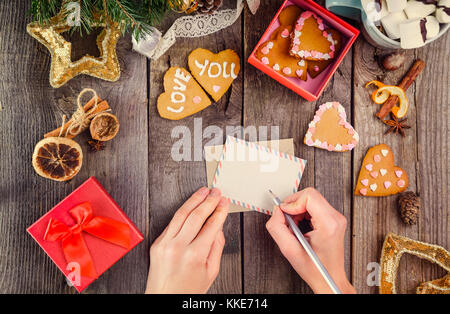 Vista superiore mani femminili desideri scrivere sul biglietto di auguri per una persona amata per natale. tavolo in legno con arredamento, scatola con biscotti fatti in casa nella forma di Foto Stock