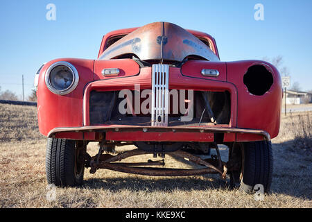 Manca la griglia anteriore e il faro anteriore prese di un vecchio abbandonato arrugginimento auto rossa in campo rurale in un basso angolo di visione Foto Stock