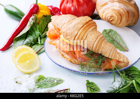 Croissant sandwich con salmone salato sulla piastra bianca, servito con insalata fresca foglie di limone, il sale di mare e le verdure sopra azzurro sfondo di legno Foto Stock