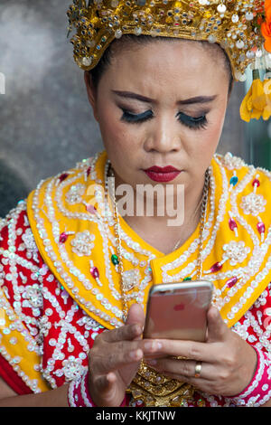 Bangkok, Tailandia. Thai danzatrice presso il Santuario di Erawan controllando il suo telefono cellulare. Foto Stock