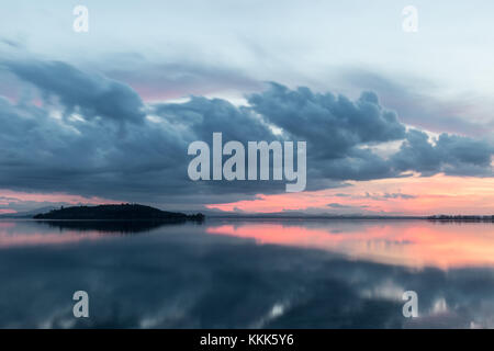 Vista di un lago al tramonto, con perfetta riflessioni di un'isola e alcuni grandi nuvole su acqua Foto Stock