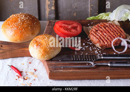 Ingredienti per fare in casa burger sul tagliere di legno, servita con carni coltello e forchetta sulla tovaglia bianca. scuro in stile rustico. Foto Stock