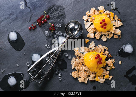 Mango sorbetto gelato, servita su wafer briciole con ribes rosso bacche, scoop di metallo e i cubetti di ghiaccio su nero ardesia. vista dall'alto. Foto Stock
