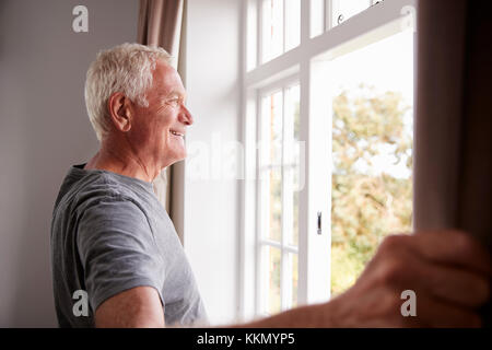 Senior uomo apertura camera da letto tende e guardando fuori della finestra Foto Stock